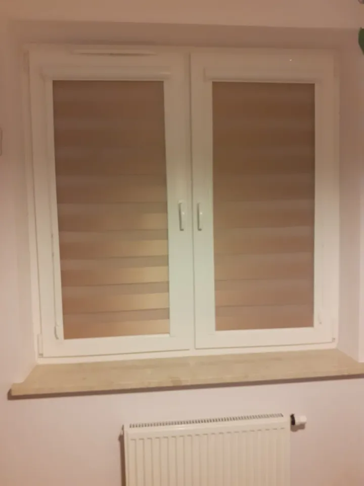Beżowe rolety dzień i noc na białym oknie dwuskrzydłowym nad grzejnikiem