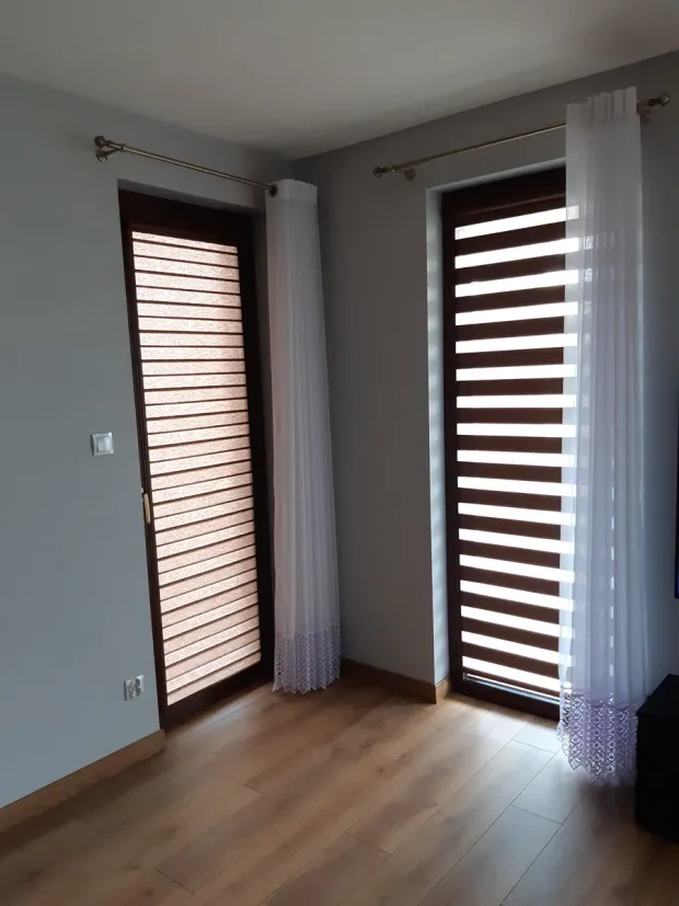 Rolety dzień i noc na wysokich oknach salonu z firanami