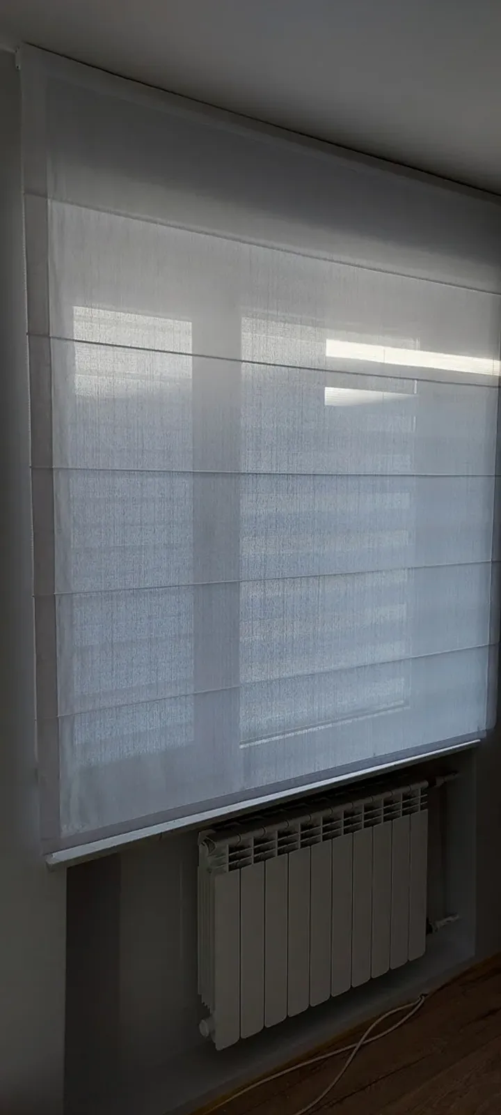 Biała roleta dzień i noc nad grzejnikiem, pasy w pozycji przepuszczającej światło